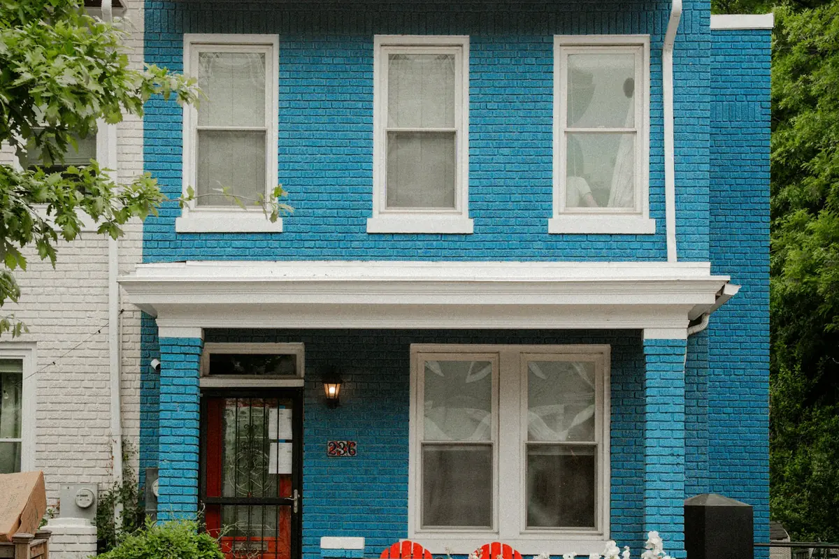 A blue brick two-story house with a red front door behind a low brick wall