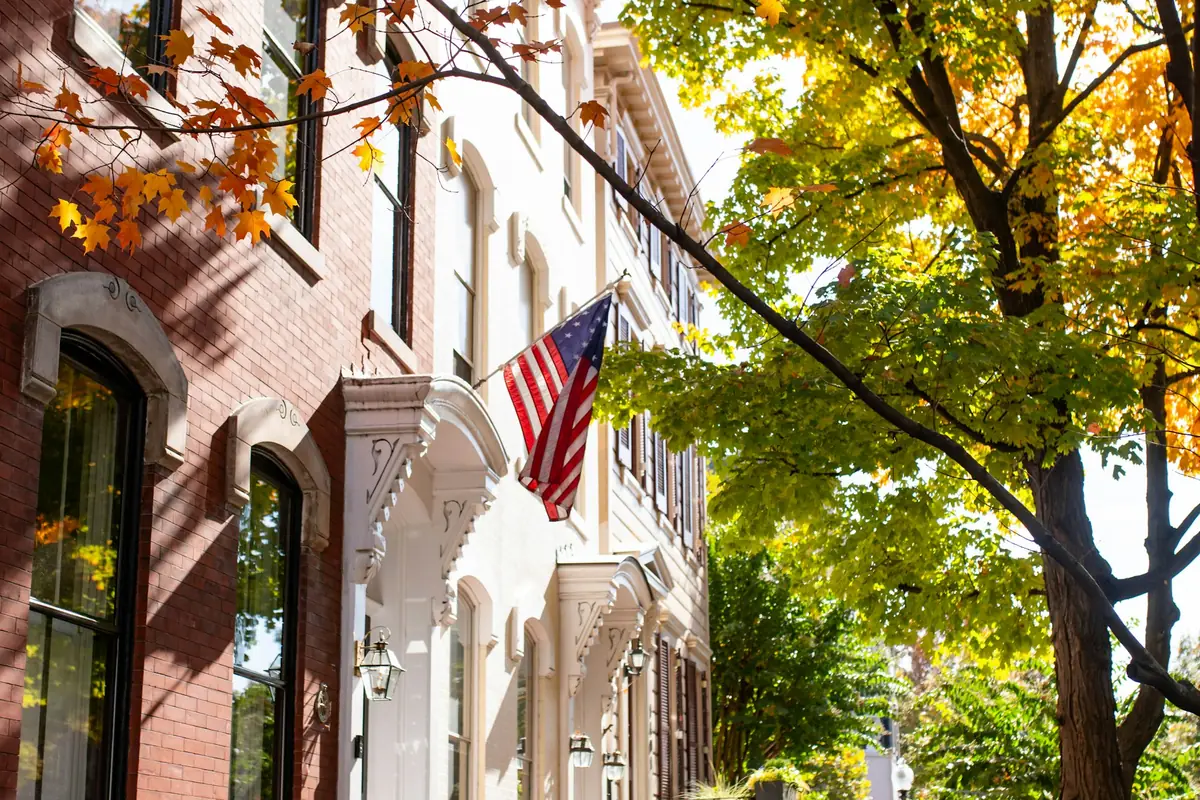 Brick rowhouses with iron stair railings and a flag, on a leafy sidewalk in fall
